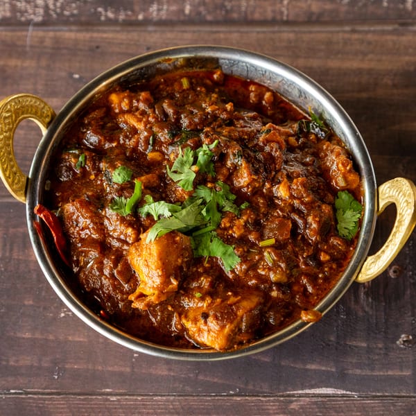 Rich, dark brown chicken curry with fresh cilantro in a silver bowl with gold handles on a wooden table.