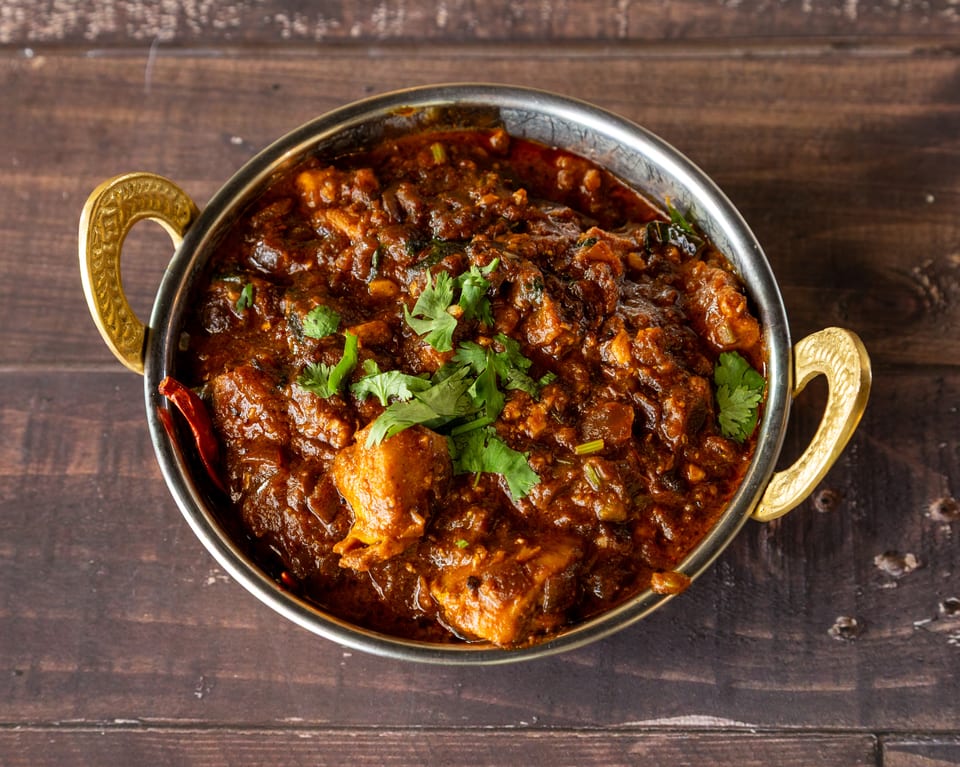 Rich, dark brown chicken curry with fresh cilantro in a silver bowl with gold handles on a wooden table.