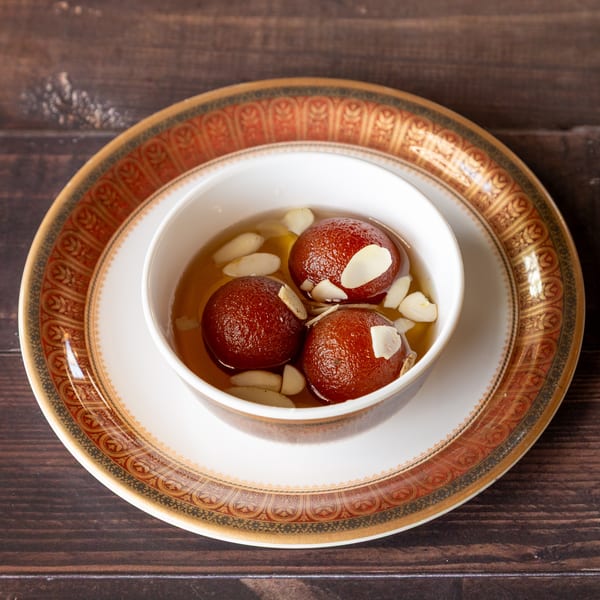 Three gulab jamun in syrup with almond slivers, served in a white bowl on a decorative plate.