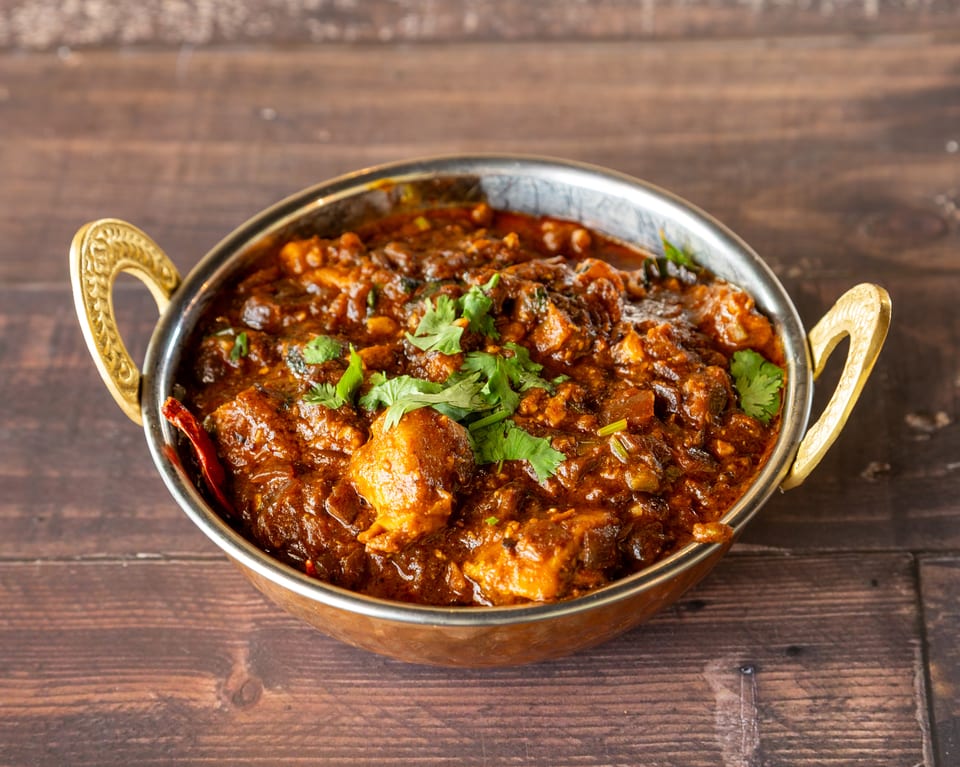 Rich, dark brown Indian curry with chicken and cilantro in a brass bowl on a wooden table.