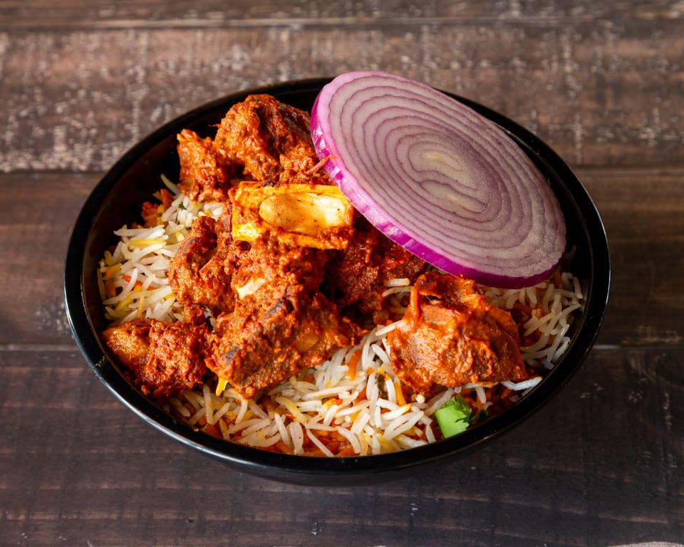 Spicy lamb biryani with basmati rice and a slice of red onion in a black bowl.