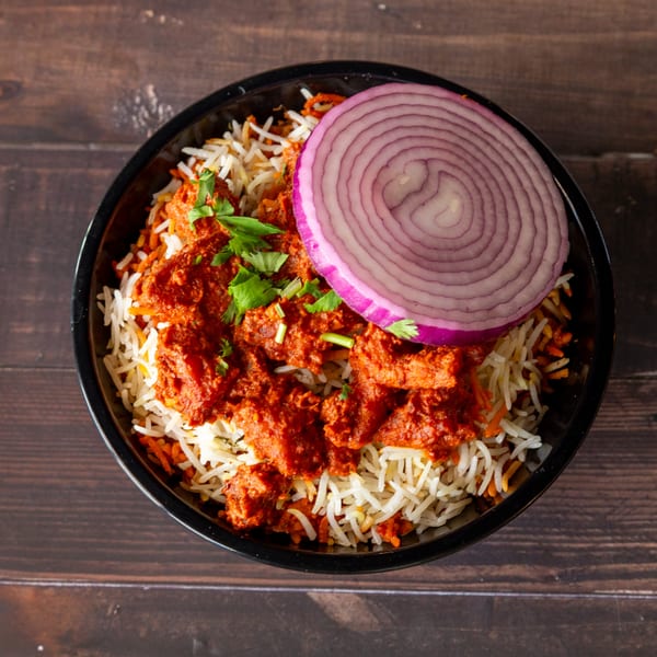 Overhead view of chicken biryani with red onion and cilantro in a black bowl on a wooden table.