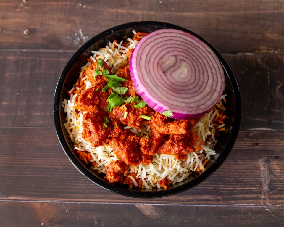 Overhead view of chicken biryani with red onion and cilantro in a black bowl on a wooden table.