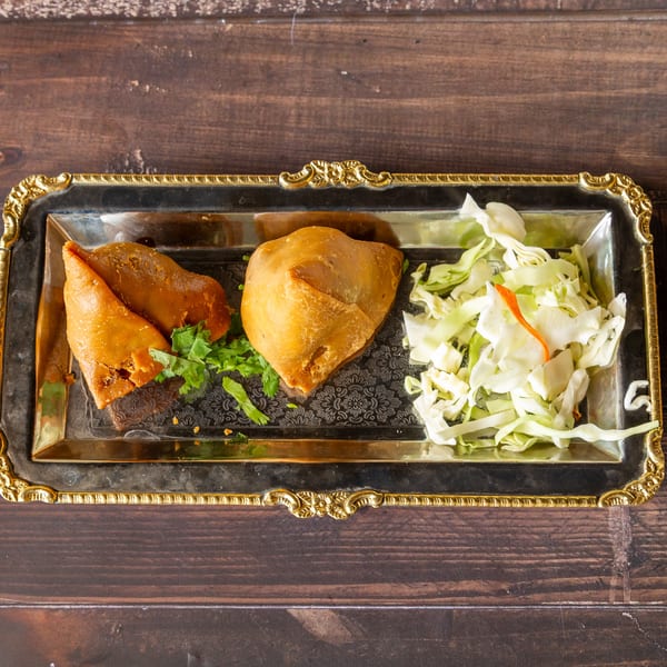 Two golden-brown samosas garnished with cilantro, served with shredded cabbage on a decorative silver tray.