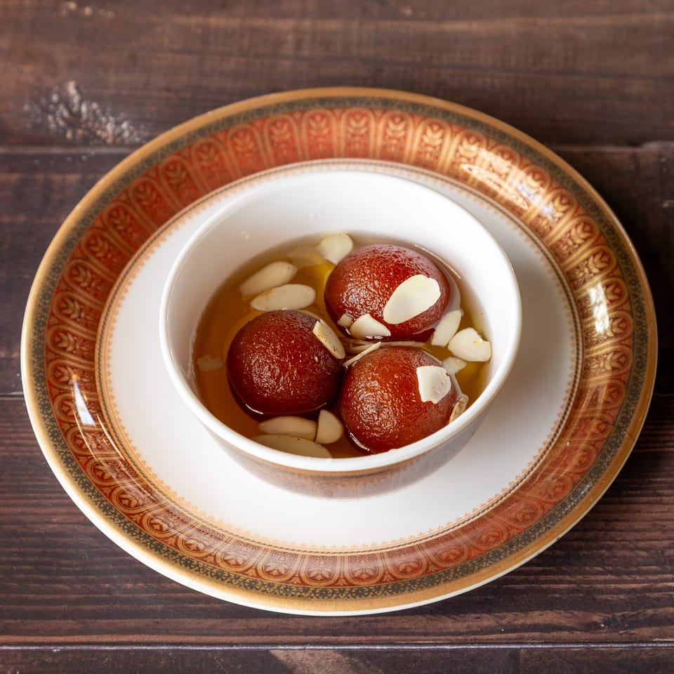 Three gulab jamun in syrup with almond slivers, served in a white bowl on an ornate plate.