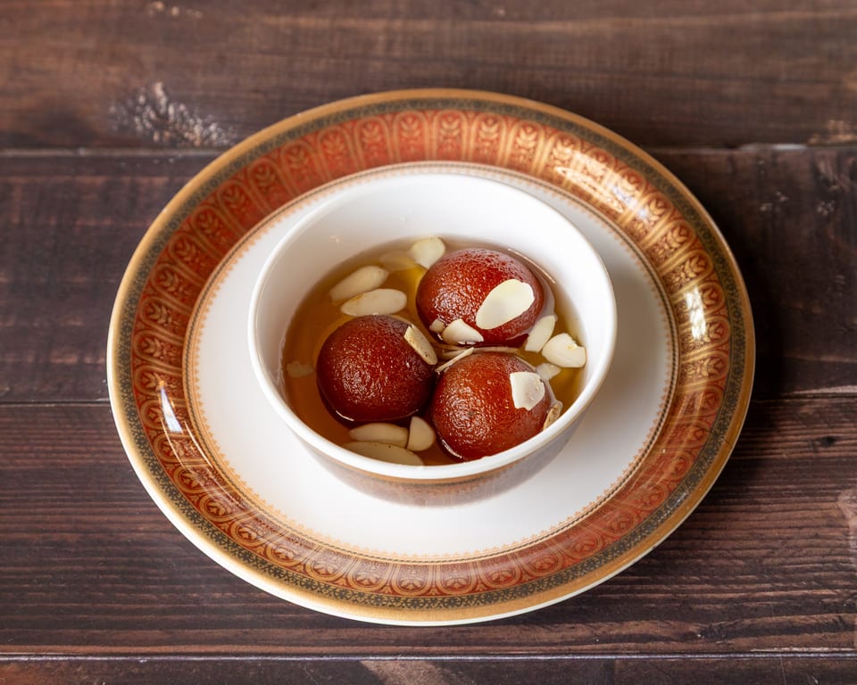 Three gulab jamun in syrup with almond slivers, served in a white bowl on an ornate plate.