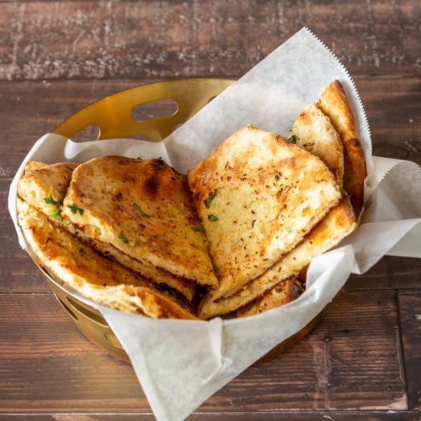 Golden brown naan bread pieces, garnished with herbs, served in a brass basket lined with parchment paper.