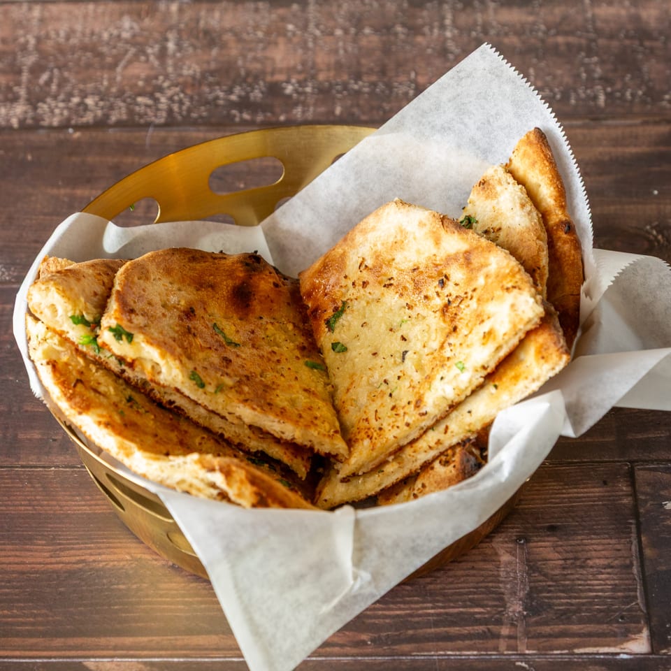 Golden brown naan bread pieces, garnished with herbs, served in a brass basket lined with parchment paper.