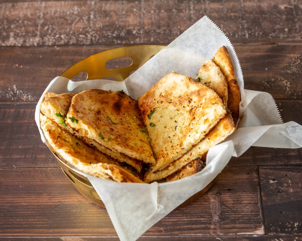 Golden brown naan bread pieces, garnished with herbs, served in a brass basket lined with parchment paper.