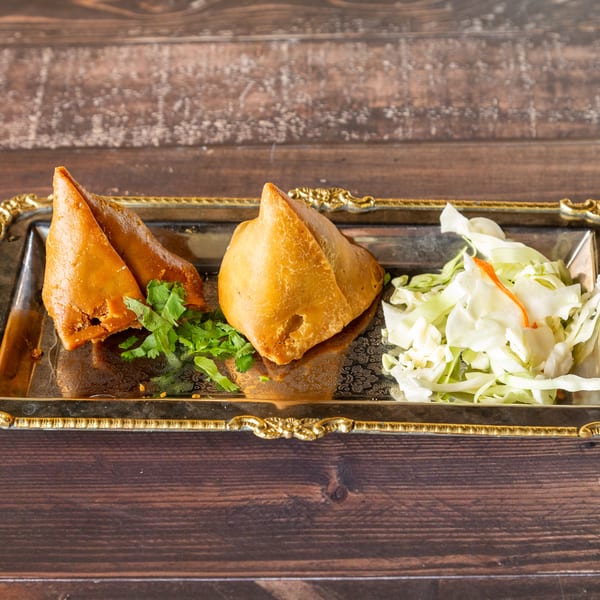 Two golden-brown samosas with cilantro and shredded cabbage on a silver and gold tray.
