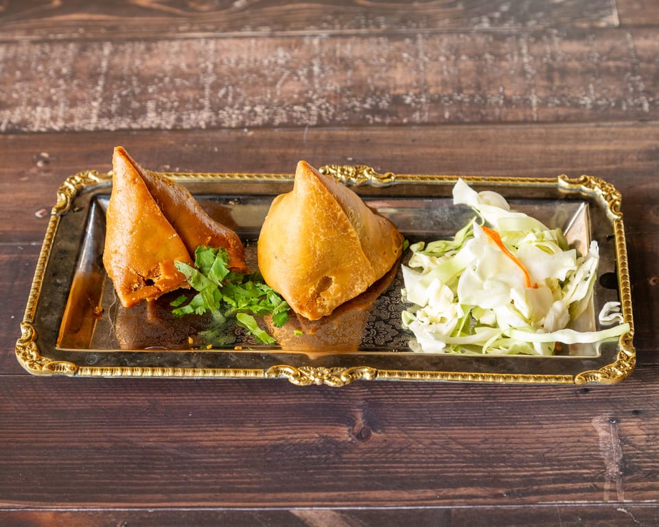 Two golden-brown samosas with cilantro and shredded cabbage on a silver and gold tray.
