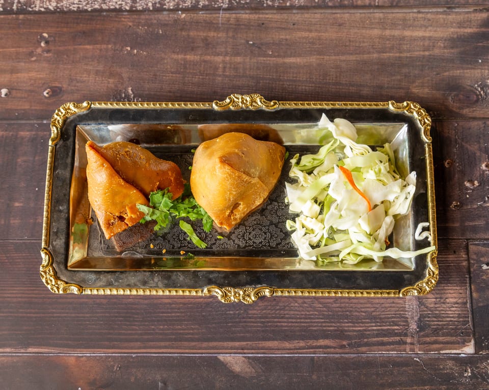 Two golden-brown samosas with cilantro and shredded cabbage on a decorative silver tray.