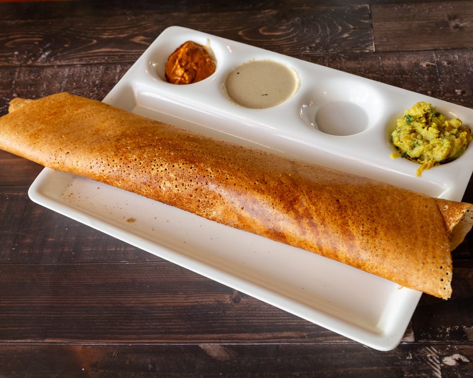 Crispy golden dosa served on a white rectangular plate with three small bowls of chutneys and potato curry.